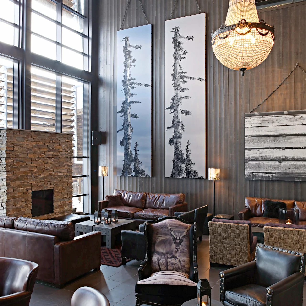 Luxurious chandelier illuminating a cozy lounge with leather sofas and armchairs, stone fireplace, large windows, and wall art of trees. Warm, sophisticated atmosphere in a modern setting.  