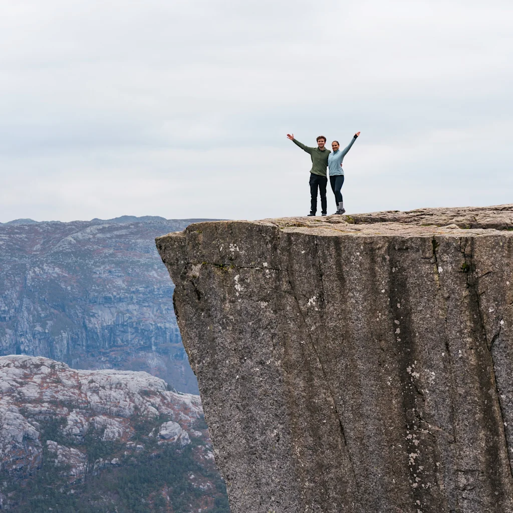 Kaksi ihmistä juhlii saavutettuaan vuorenkallion reunan, kädet kohotettuina ilosta, näkymä laajaan, pilviseen maisemaan.