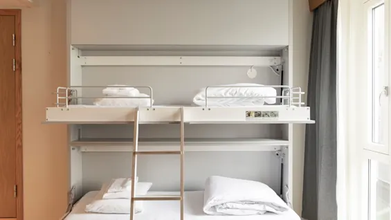 A clean, modern bunk bed with white bedding and a ladder, set in a bright room with a window and grey curtain.