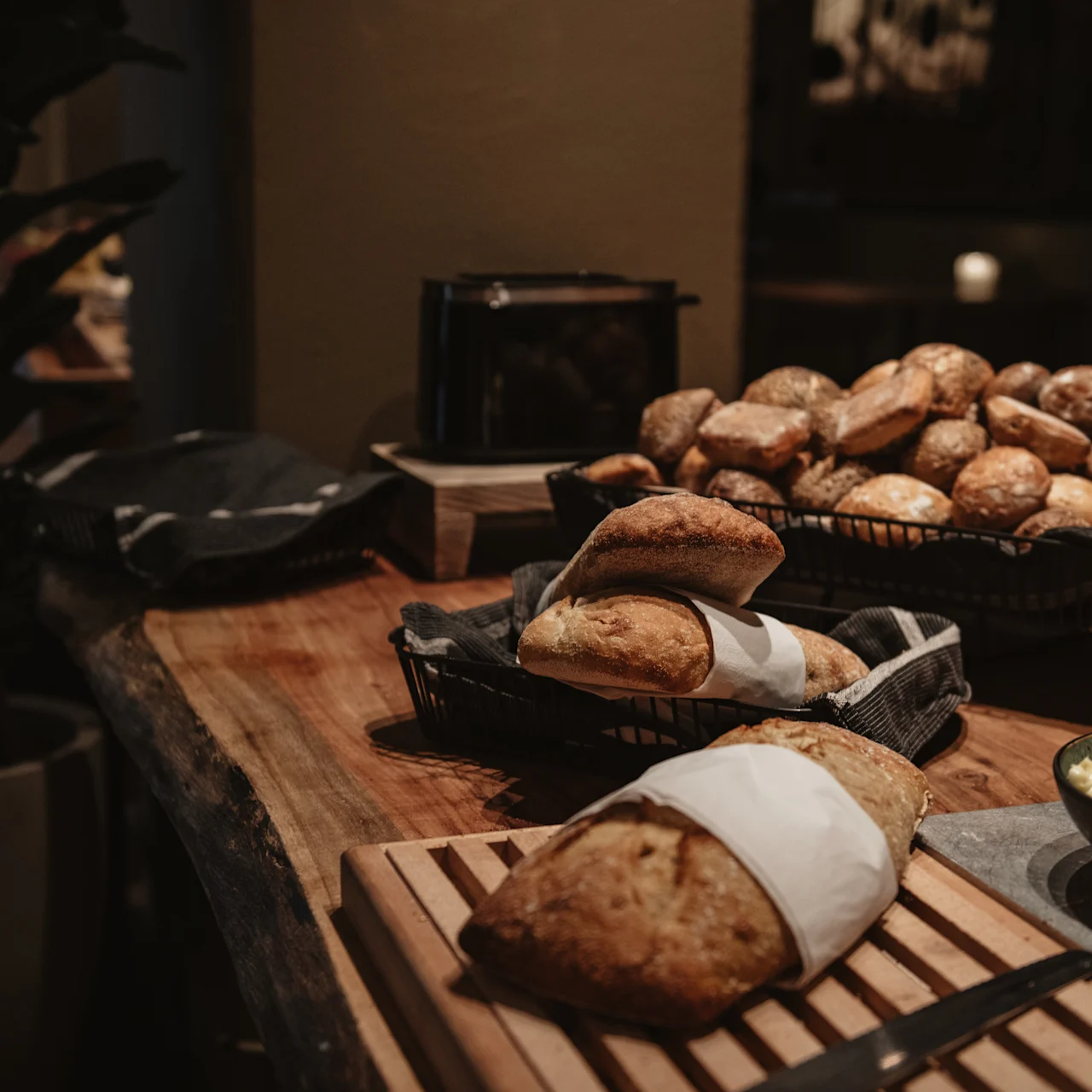 Bread sits on a wooden table surrounded by baskets filled with various rolls. Butter is placed in a bowl to the side. The setting appears to be a cozy dining area.