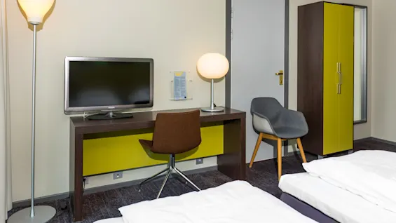 A hotel room features a desk with a lamp and a Philips TV, a brown chair, adjacent modern gray chair, and a wardrobe near a closed door.