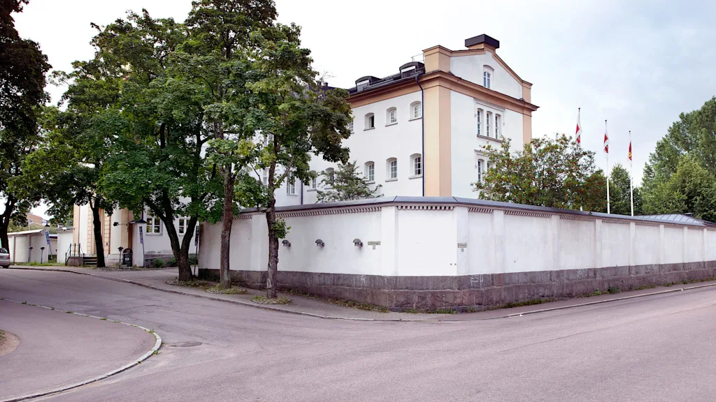 A historic building at a street corner, surrounded by a high wall, with a tree-lined sidewalk and multiple national flags flying.