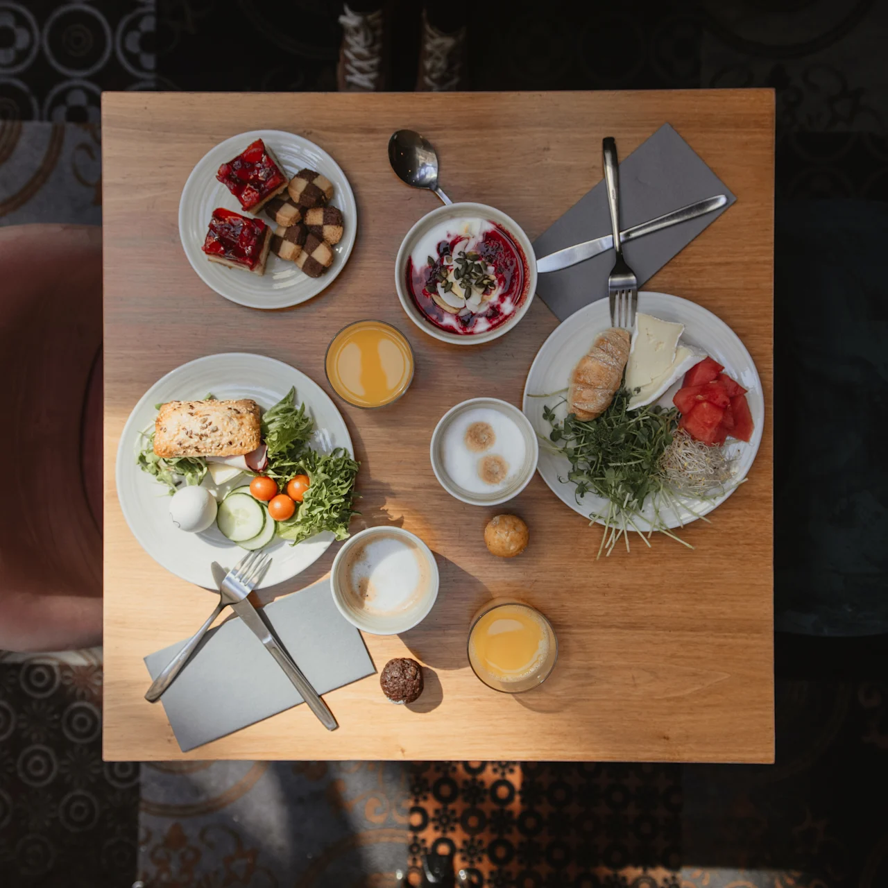 Morgenmadsretter ligger på et træbord og viser bagværk, frugt og salater med appelsinjuice og kaffe. En lyserød stol og mørkt mønstret gulv omkranser.