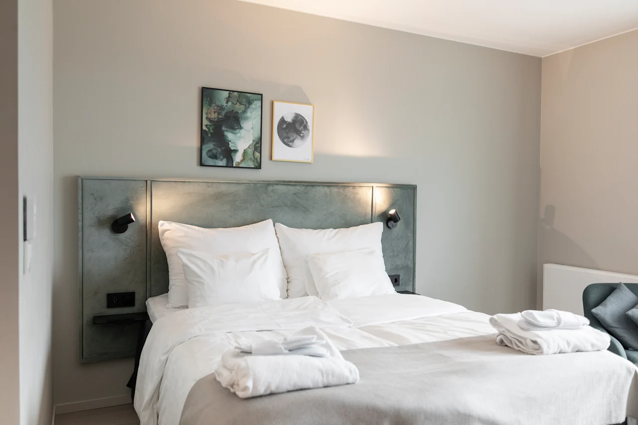 Viihtyisä hotellihuone, jossa siististi pedattu sänky, valkoiset liinavaatteet ja kaksi taideteosta sängynpäädyn yläpuolella. Puhtaat pyyhkeet ovat valmiina.