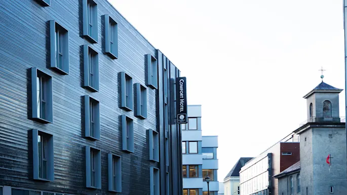 Moderni rakennus, jossa on ulkonevat ikkunat, ja kyltti "COMFORT HOTEL" kaupunkikadulla. Vieressä on pieni, perinteinen kirkontorni ristillä.