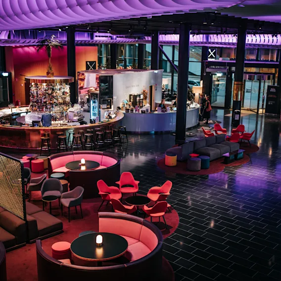En rund bar med pallar inhyser besökare i en modern lounge med färgglada stolar och soffor. Mjukt lila och blått ljus skapar en livlig, inbjudande atmosfär.