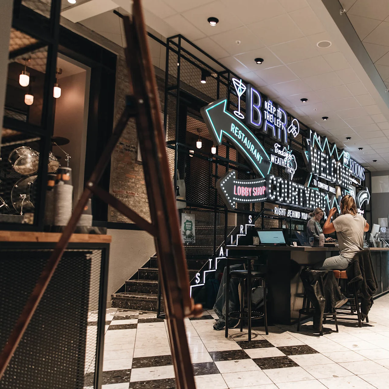 En person sitter ved en bardisk med en laptop, i et moderne interiør med lyse neonskilt som viser "BAR," "RESTAURANT," "LOBBY SHOP" og forskjellige retninger.