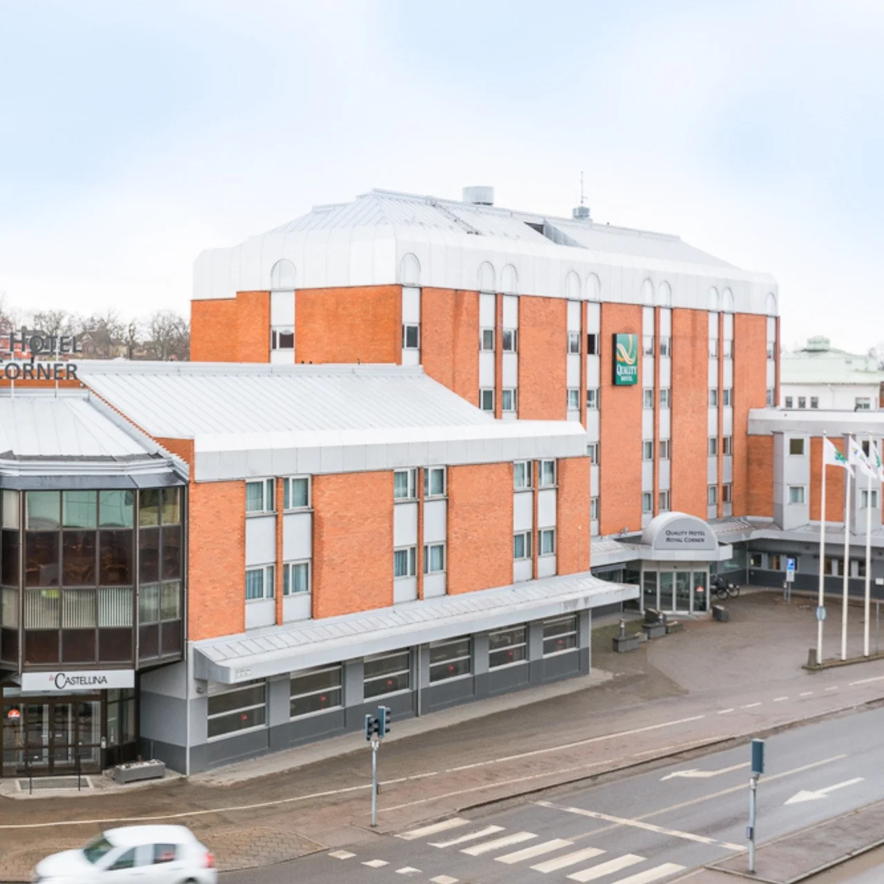 A multi-story brick and glass hotel building stands on a city street corner. Four flags fly nearby. Signage reads: “QUALITY HOTEL ROYAL CORNER” and “OBJEKTIA”.