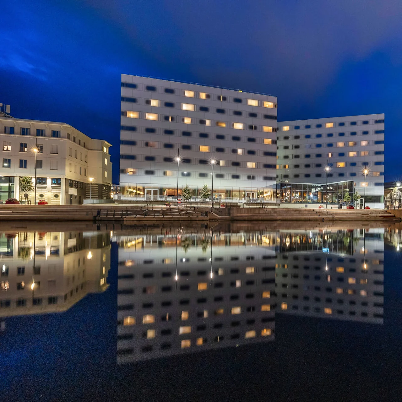 A modern building with illuminated windows reflects in the calm water at night, surrounded by other structures and streetlights on a waterfront. 