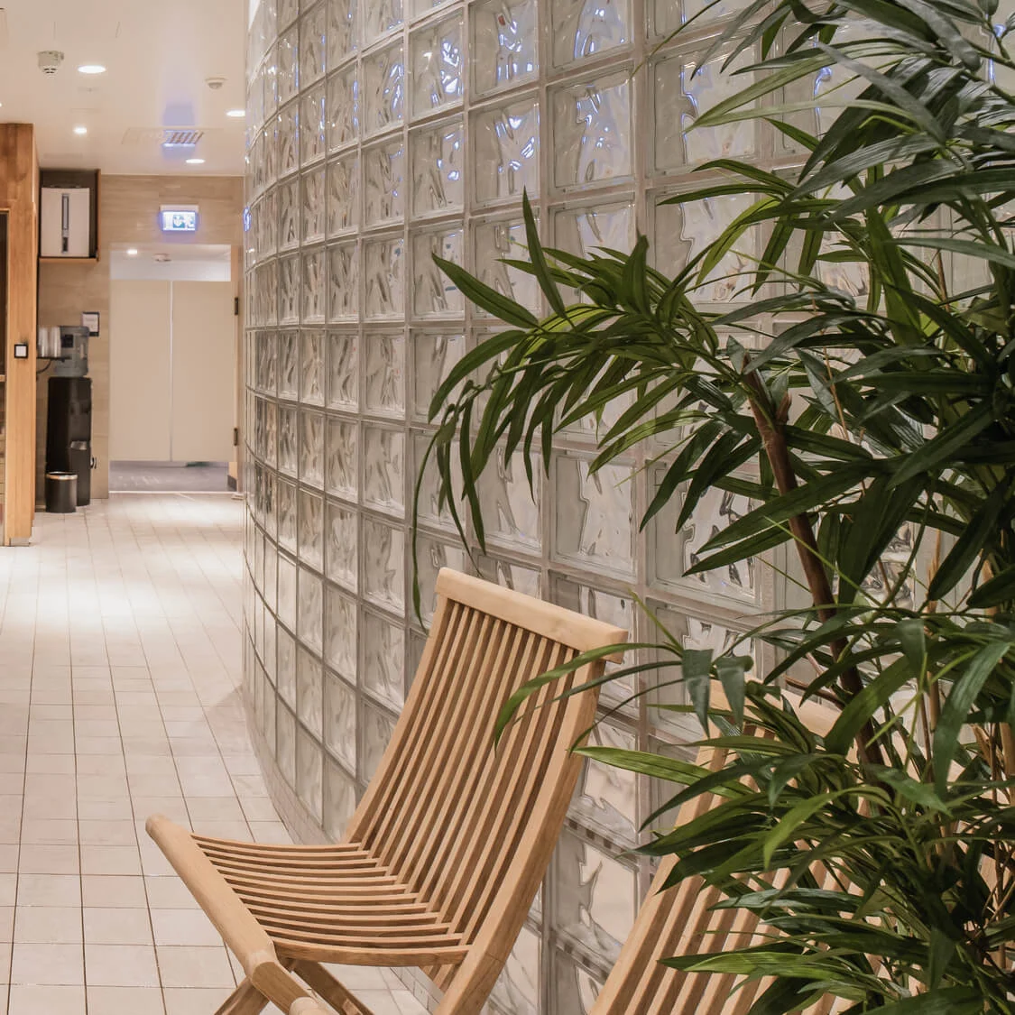 A wooden sauna door is adjacent to a tiled hot tub surrounded by plants, a wooden chair, and glass block walls. Towels are stacked nearby on a shelf.
