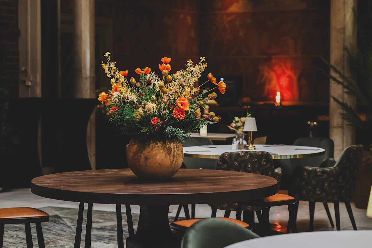 A large bouquet of vibrant flowers sits in a round vase on a wooden table, surrounded by cushioned chairs in a softly lit, elegant dining area.