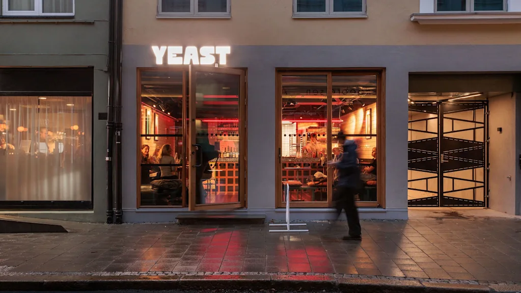 A vibrant restaurant Yeast at dusk with people dining inside, and a person walking on the wet sidewalk outside.