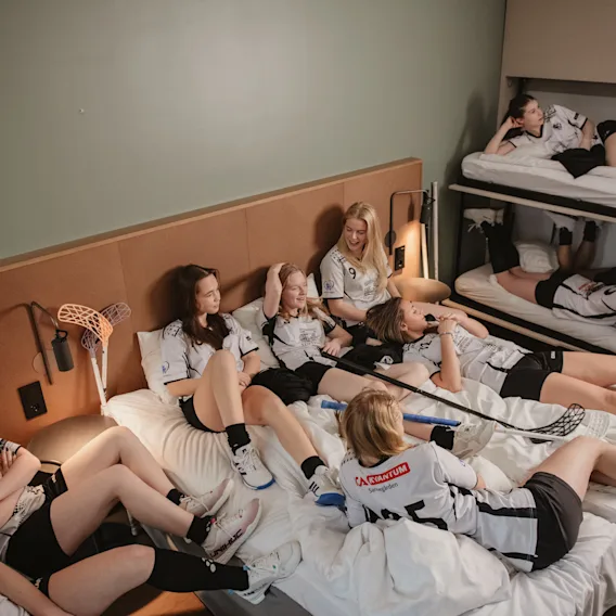 En gruppe unge idrettsutøvere slapper av på et hotellrom, noen på senger og andre i køyesenger, etter en kamp eller trening.