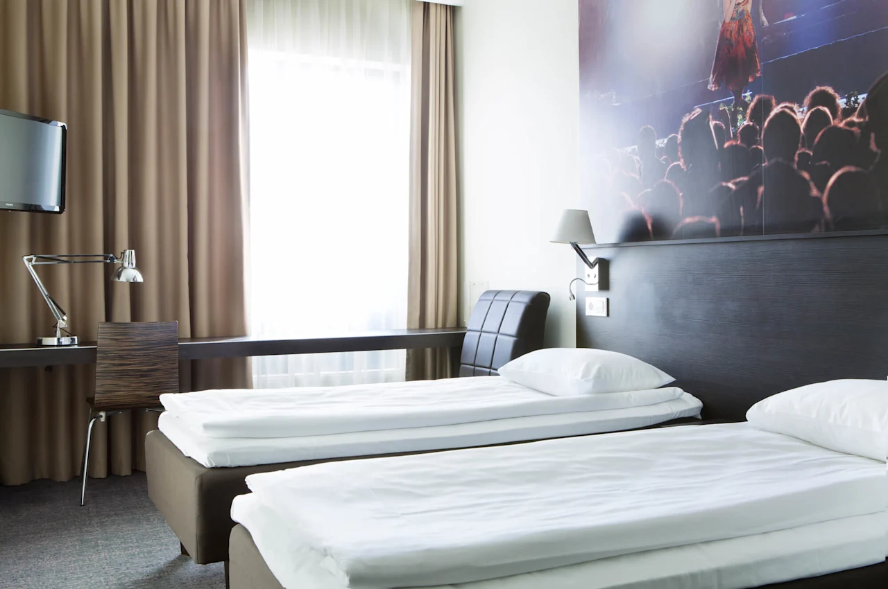 Two neatly made single beds with white linens stand in a modern hotel room. A desk with a lamp and chair sits beside a window with beige curtains.