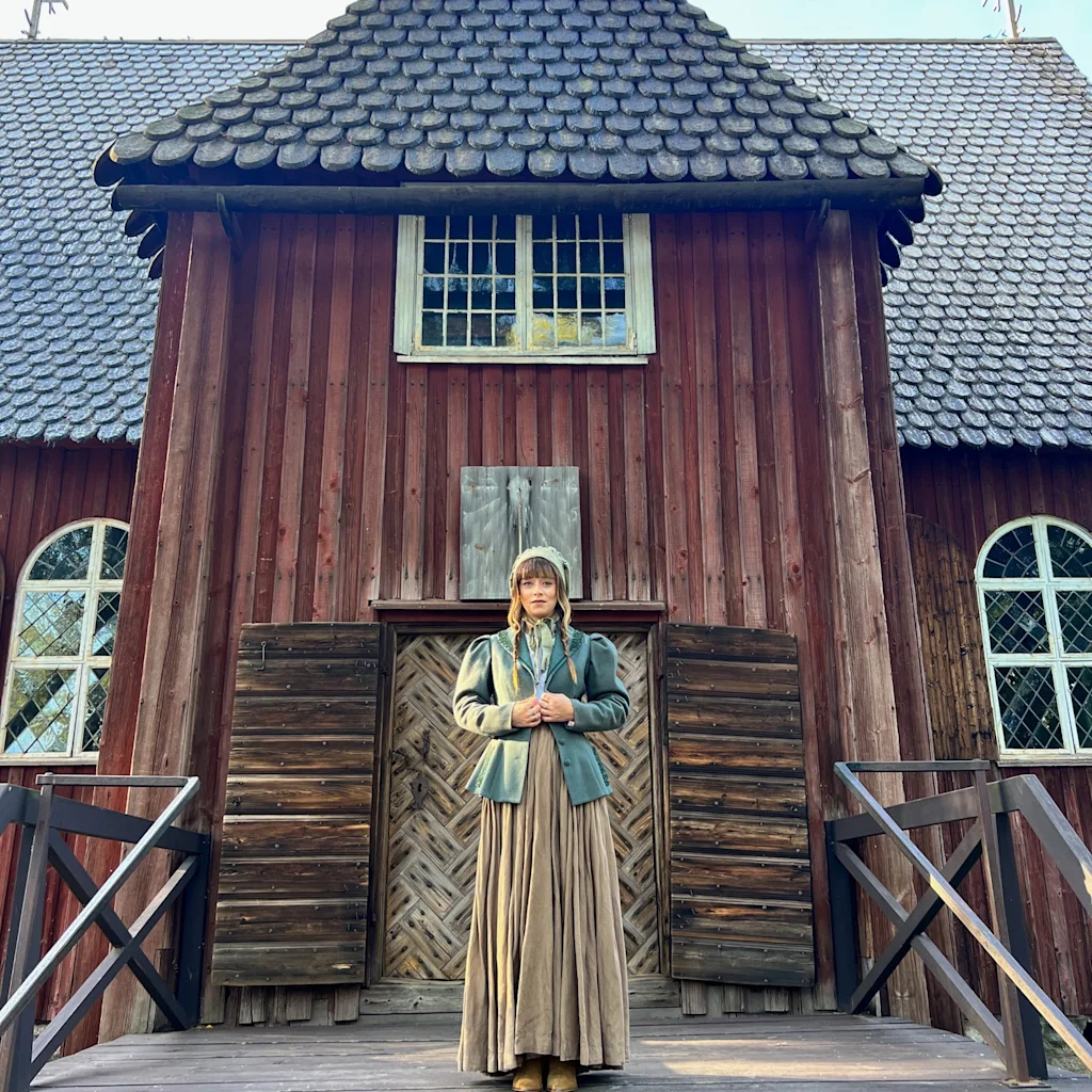 En person i historisk antrekk står på en veranda foran en rød trebygning ved Seurasaari friluftsmuseum.