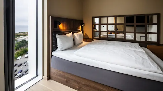 Et koselig hotellrom med en pent oppredd seng med hvitt sengetøy, belysning ved sengegavlen, og en stor speilvegg som reflekterer det moderne interiøret ved siden av et høyt vindu med utsikt.