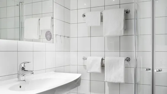Ett handfat med en kromkran står mot vita kakelväggar i ett rent badrum. Vita handdukar hänger på stänger. En spegel reflekterar en del av rummet.