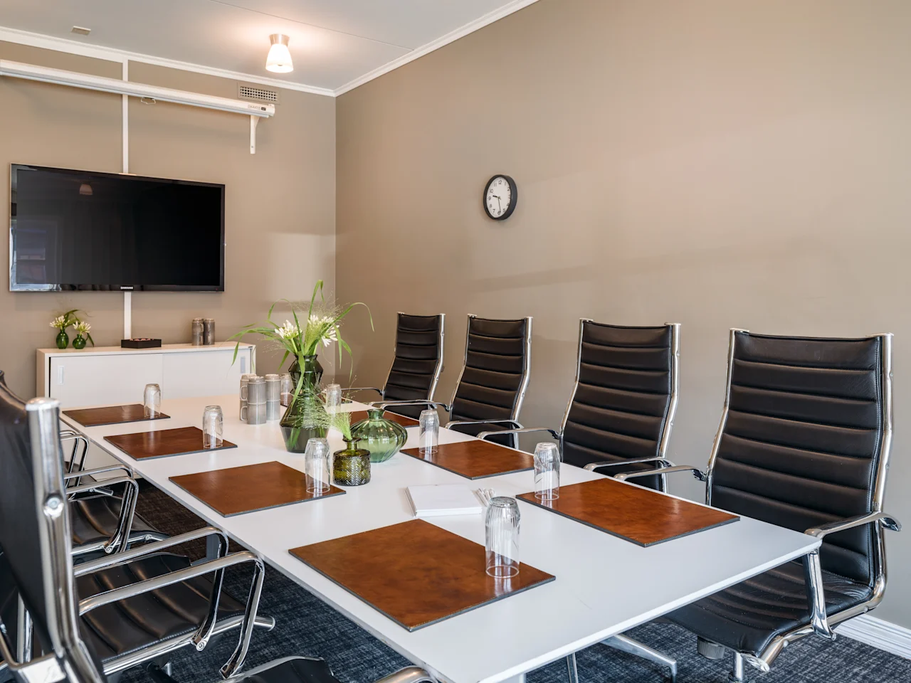 Ett modernt konferensrum med ett långt bord, svarta stolar, en TV-skärm och dekorativa växter, redo för ett möte.