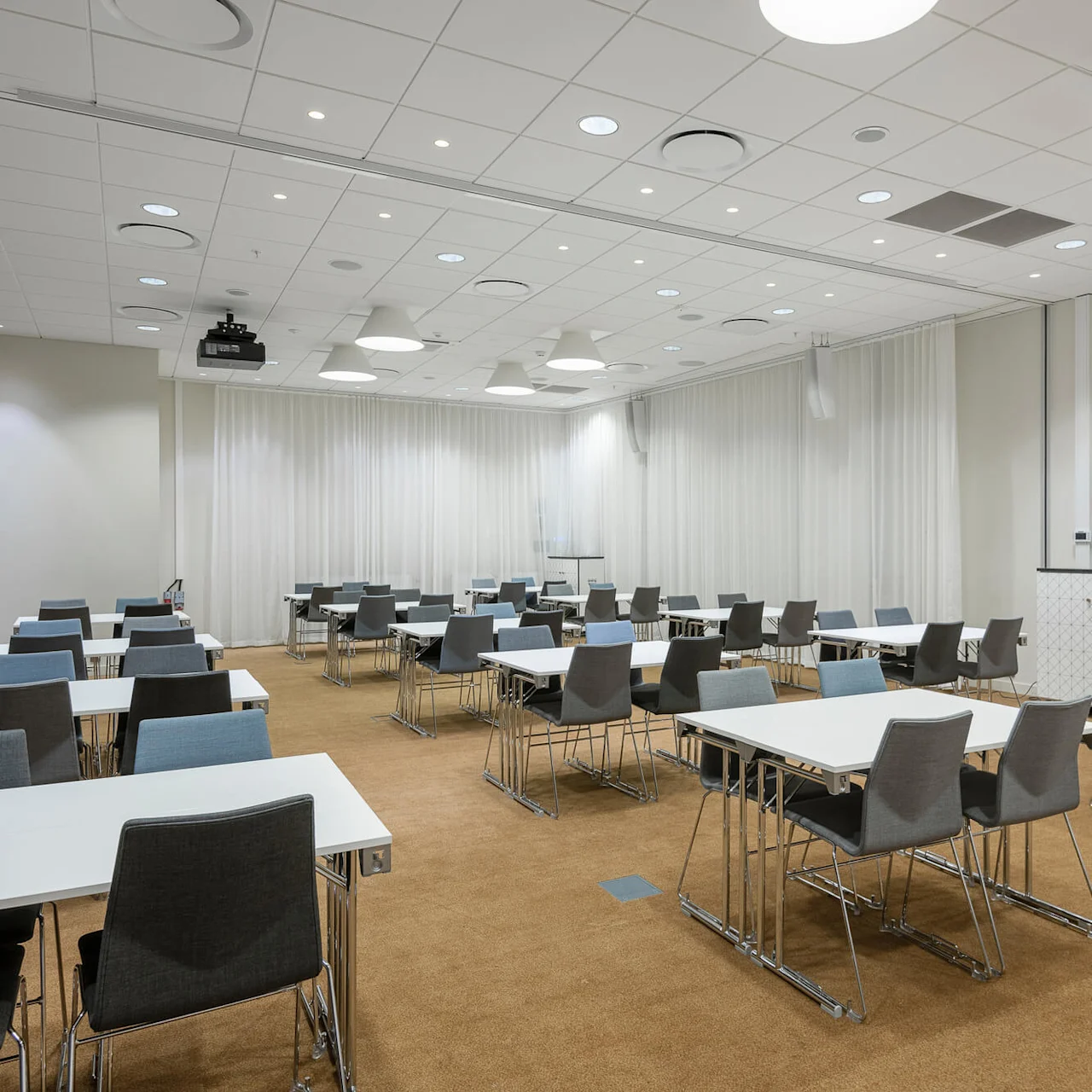 Tables and chairs are neatly arranged in rows in a bright conference room with white walls, curtains, and a beige carpet floor. Ceiling lights provide illumination.
