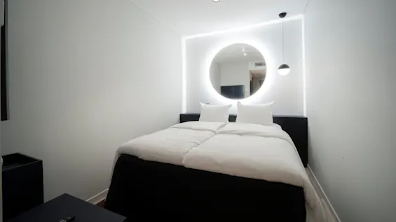 A bed, neatly made, rests beneath a large circular mirror illuminated by LED lights on a monochromatic wall. A hanging lamp is beside it in a minimalist bedroom.
