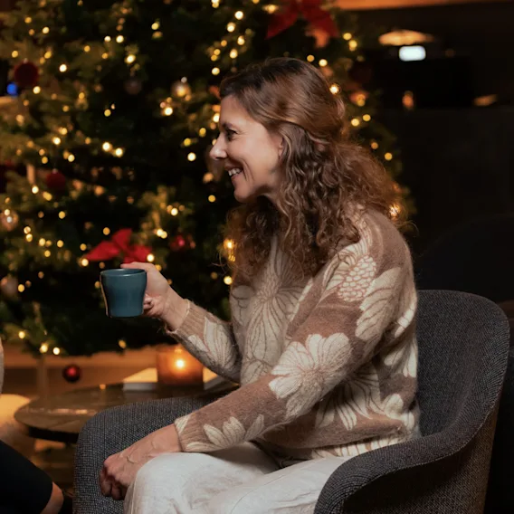 En kvinne i en koselig strikkegenser sitter med en kopp og smiler ved siden av et glitrende juletre.