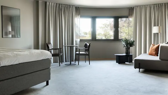 A spacious, modern hotel room features a bed, chairs by a round table, and a sofa with cushions. Large windows provide natural light, overlooking a leafy exterior.