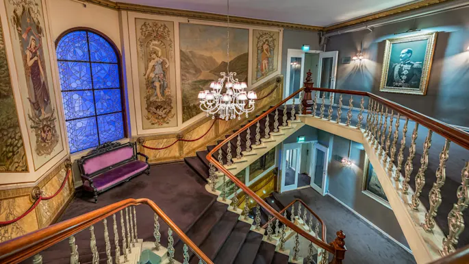 Grand staircase curves upward, adorned with ornate paintings and a central chandelier, set in a luxurious, art-filled interior featuring a purple bench and framed portrait on the wall.