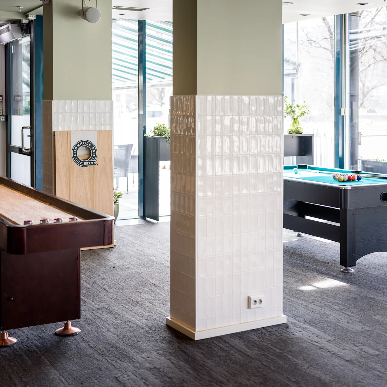 Shuffleboard and foosball tables stand idle in a bright, modern recreation room with large windows, sunlight streaming in, and greenery outside. The room features tiled pillars and a carpeted floor.