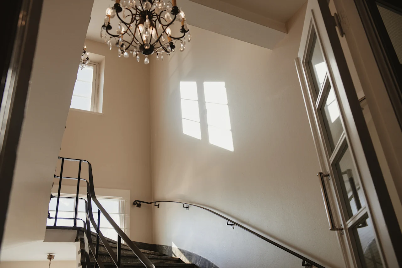 En ljus inre trappa med en ljuskrona och solljus som strömmar in genom ett fönster och kastar mönster på väggen.