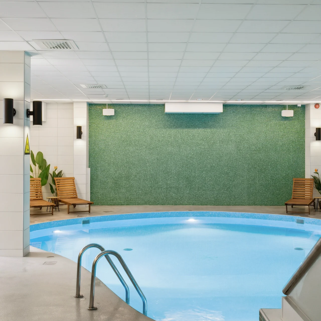 En lille, oplyst indendørs pool reflekterer blåt vand, omgivet af flisevægge, liggestole og potteplanter, hvilket skaber en afslappende fritidssammensætning.