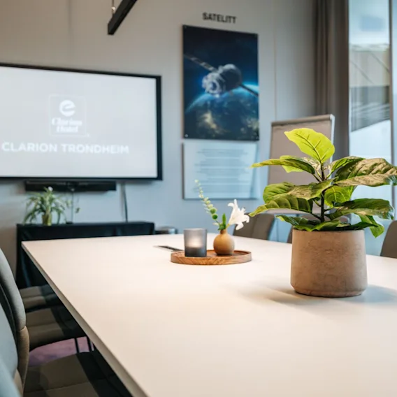 Et konferanserom med et bord som viser en plante, lys og vase. En skjerm med teksten “Clarion Hotel Trondheim.” En plakat merket “SATELLIT” viser et satellittbilde. Stoler langs bordet.