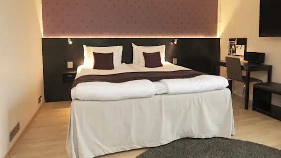 En seng med hvidt sengetøj og brune puder står i et moderne soveværelse med et mønstret sengegavl og et skrivebord med lampe og stol ved siden af.