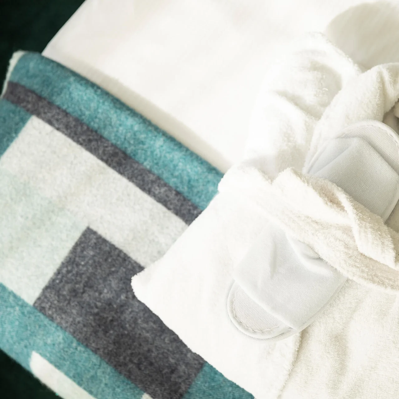 En mjuk vit morgonrock och matchande tofflor ligger prydligt på en bäddad säng, dekorerad med en geometriskt mönstrad blå och grå filt.