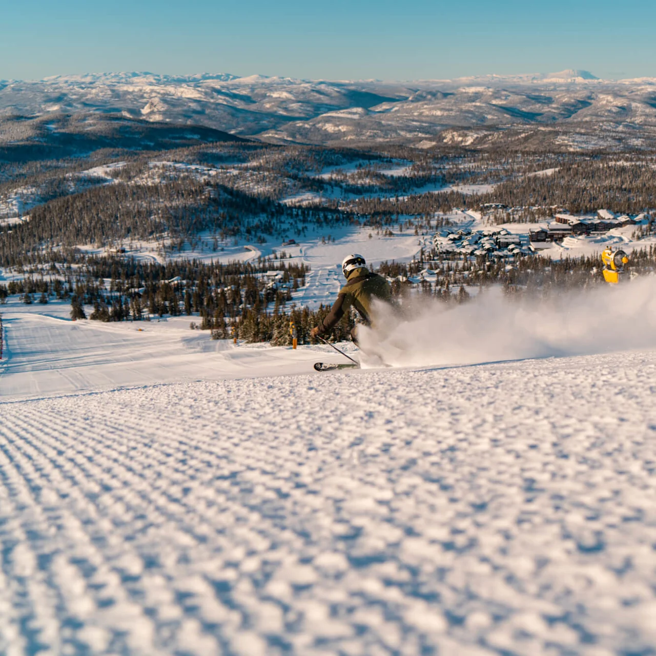 A skier speeds down a snowy slope, leaving a trail of powder behind. Snow-covered mountains and a village stretch across the horizon under a clear blue sky.