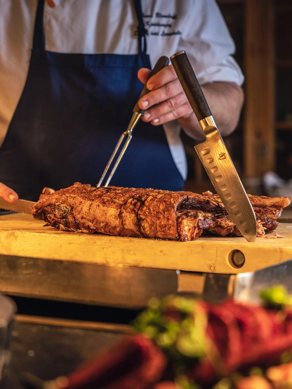 Kokk skærer opp stekt kjøtt på en skjærefjøl med kniv og gaffel. Den omkringliggende konteksten er et varmt, rustikt kjøkken med retter i forgrunnen og dempet belysning.