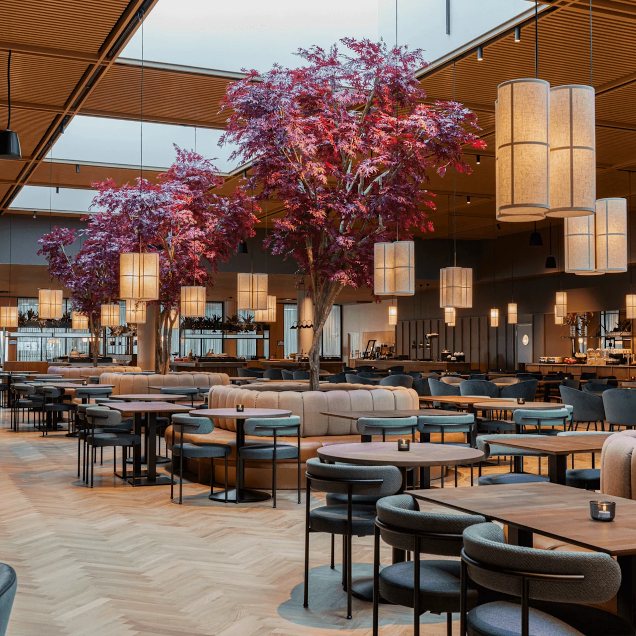 Spacious dining area with round tables and modern chairs, featuring large artificial trees with red leaves and soft lighting, creating an inviting atmosphere for guests.