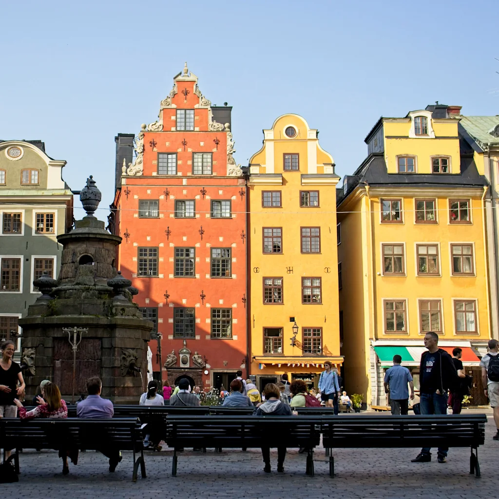 Människor kopplar av och promenerar på ett livligt torg i Gamla stan i Stockholm, omgivna av färgglada historiska byggnader.