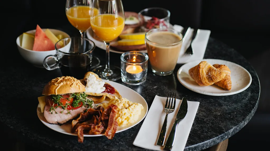 En väl presenterad frukost på ett mörkt bord inkluderar croissanter, kaffe, apelsinjuice, bacon, äggröra, bröd och frukt, med ett ljus för atmosfär.