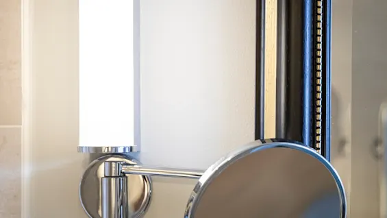 En väggmonterad, cylindrisk lampa lyser upp ett badrum med en rund spegel under, bredvid en stor spegel med ornerad ram.