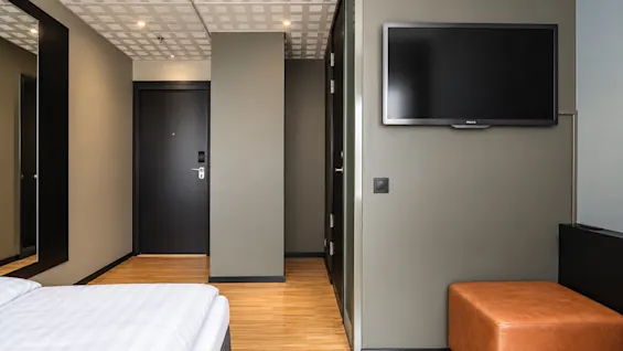 Modern hotel room featuring wooden flooring, a wall-mounted TV, a bed, and a brown ottoman.