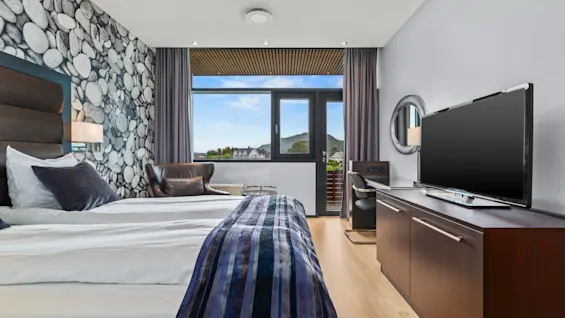 Ett modernt hotellrum med en bekväm säng, en TV och en balkong med naturskön utsikt över berg och hus.