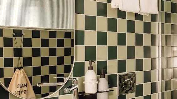 A modern bathroom at hobo oslo with green and black checkered tiles, a round mirror, sink, and towels. Lotion, soap, and hair products are visible.