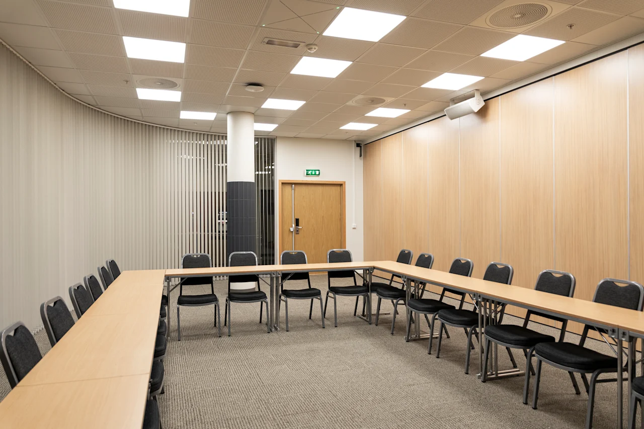 A spacious and well-lit conference room featuring a U-shaped table arrangement, light wood walls, and an exit sign.