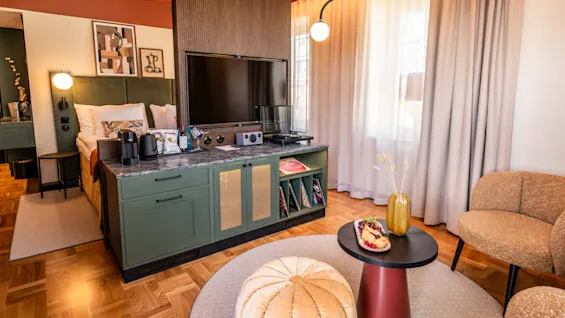 A cozy hotel room with a neatly arranged bed, large TV on a cabinet, armchair, and a table with fruit. Ambient lighting and modern decor create a warm, inviting atmosphere.