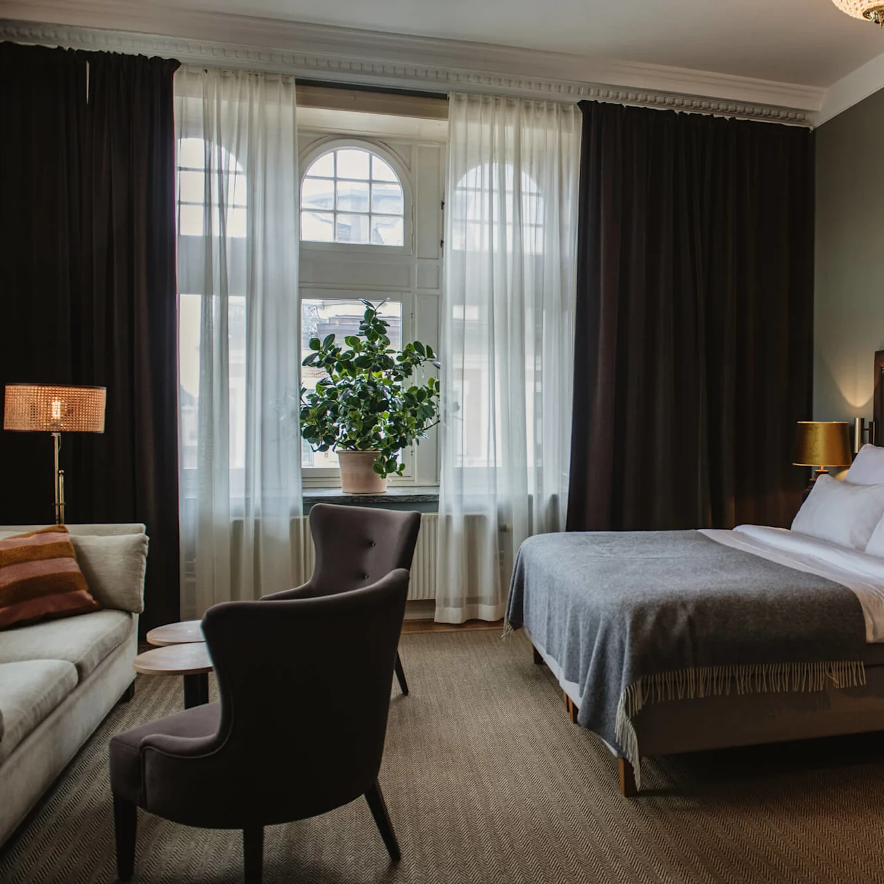A bed with white linens and a gray blanket is flanked by lamps on wooden nightstands; an armchair and sofa sit nearby, with a large window and curtains in the background.  