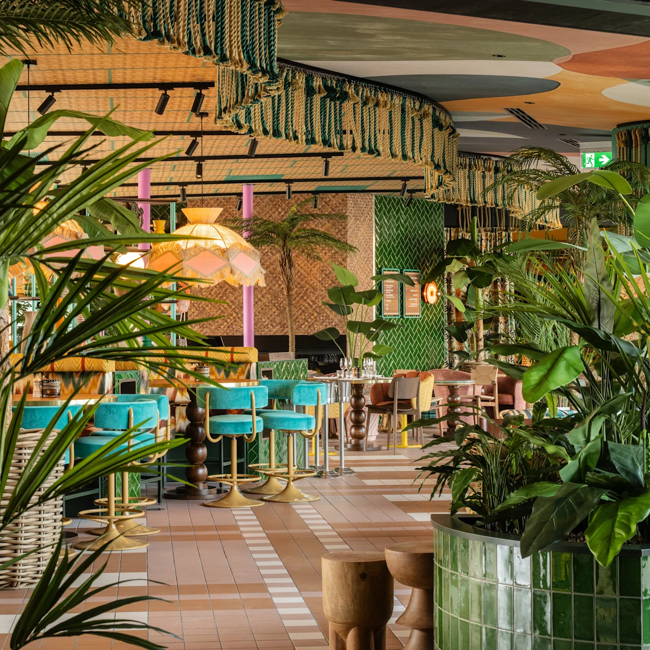 Lush greenery fills a restaurant with colorful seating and hanging fabric decor. Green tiles, vibrant furniture, and tropical plants create a vivid, inviting atmosphere.