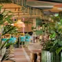 Frodig vegetation fylder en restaurant med farverige siddepladser og hængende stofdekorationer. Grønne fliser, livlige møbler og tropiske planter skaber en livlig, indbydende atmosfære.