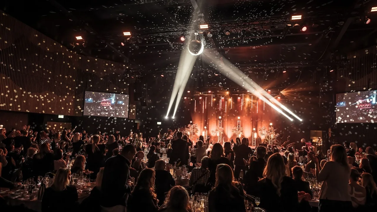 A large group of people sits and stands in a dark event venue, illuminated by bright stage lights, and surrounded by floating confetti.