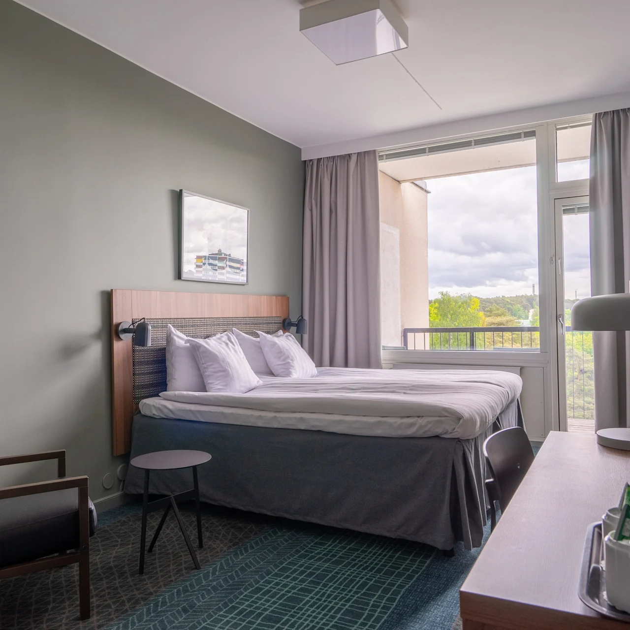 Seng er pent oppredd i et moderne hotellrom. En lenestol står ved et lite bord. Gardindekket vindu gir utsikt til utendørs. Et skrivebord har en lampe og et brett med fasiliteter.