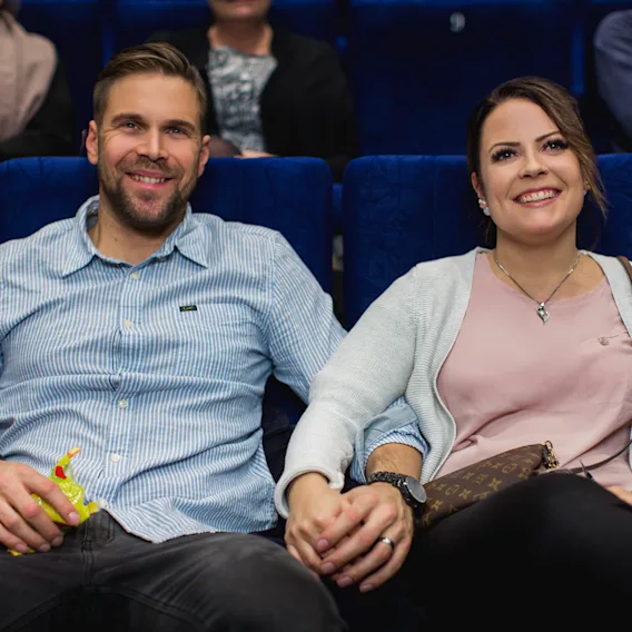Hymyilevä pariskunta pitelee toisiaan kädestä elokuvateatterissa tai auditoriossa.